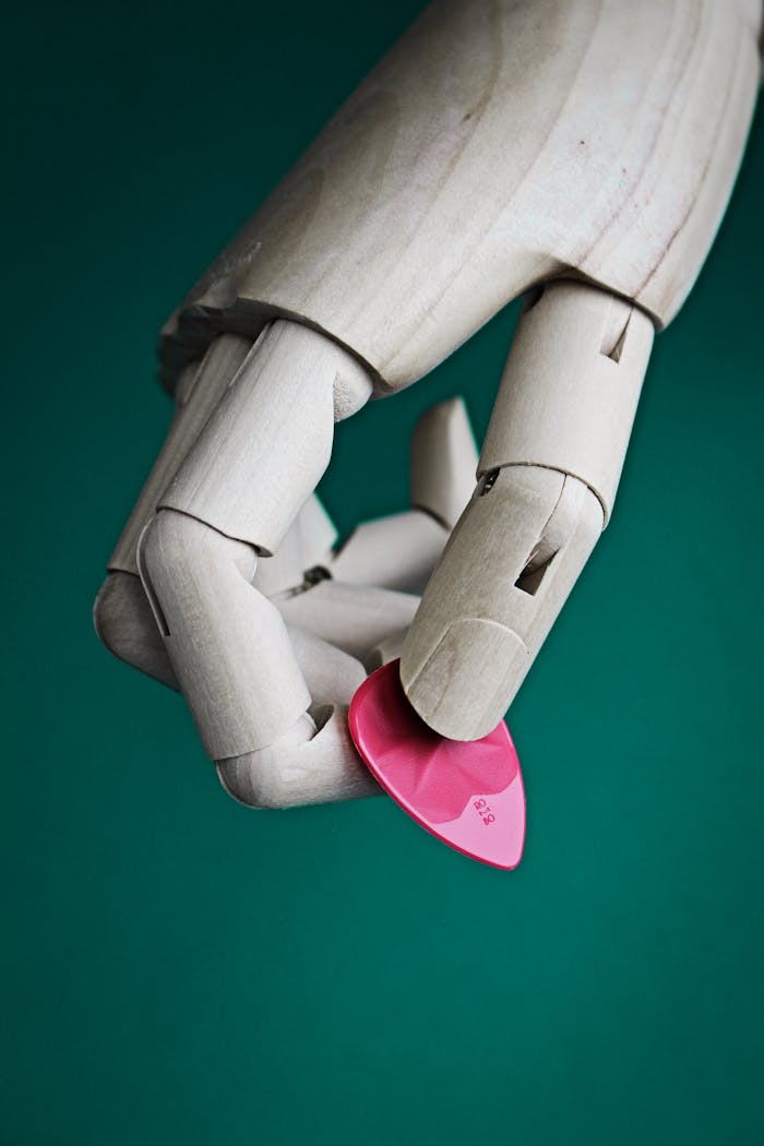 Home Close-up of a wooden mannequin hand holding a pink guitar pick against a green background.